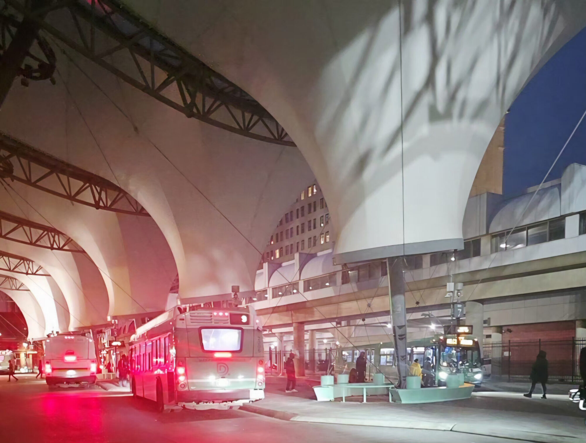 Buses idling under the Rosa Parks Transit Center canopy at night, red brake lights glowing